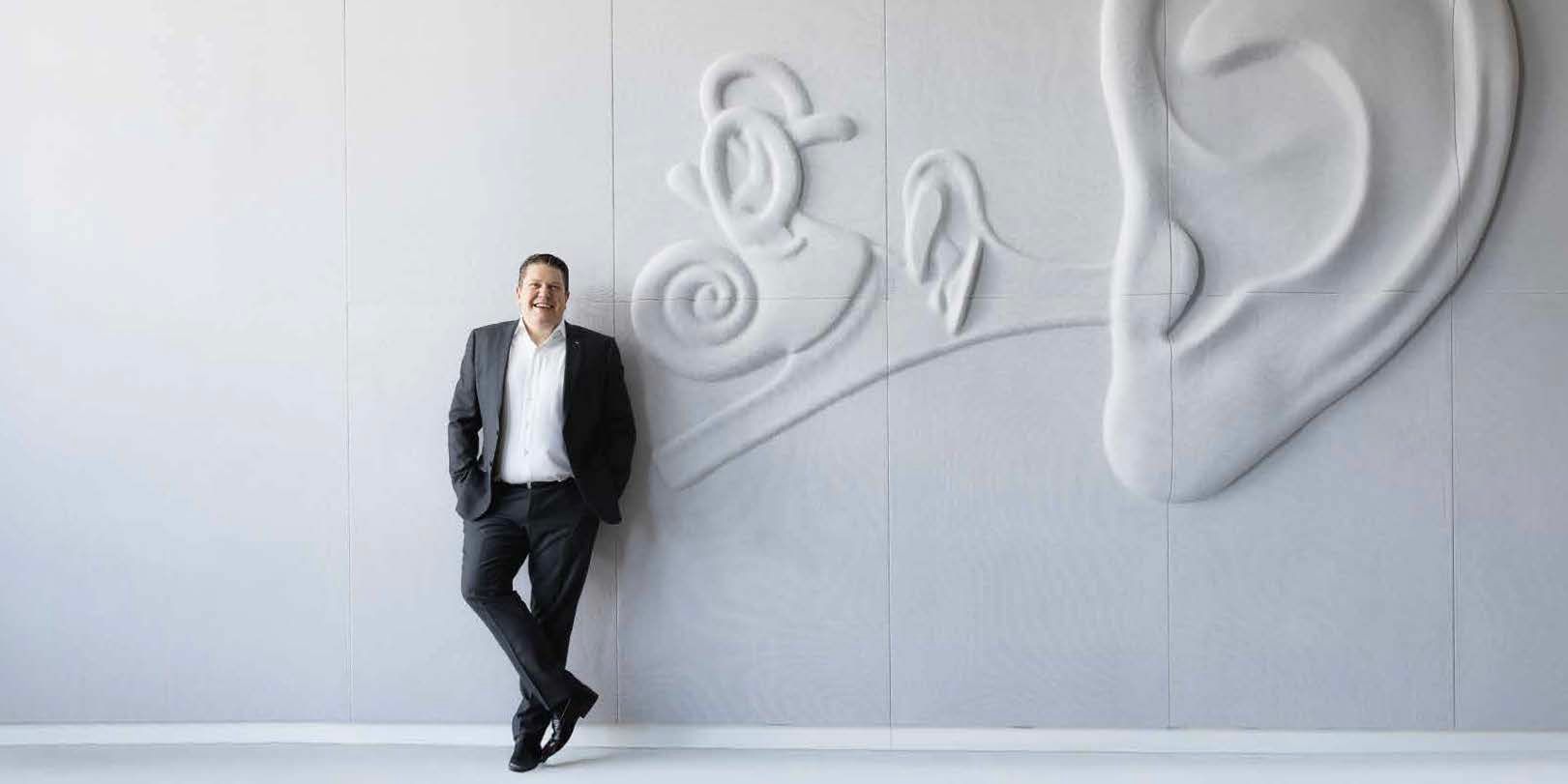 Lukas Schinko lehnt an einer grauen Wand auf der ein Ohr zu sehen ist. Er schaut in die Kamera und lächelt.